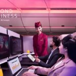 Una azafata, con el uniforme de Qatar Airways, sonríe a una mujer y a un hombre que ocupan cada uno un puesto en primera clase, en una cabina de avión.