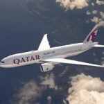 Imagen en contrapicado de un avión blanco, con el logo de Qatar Airways en la cola, que surca los aíres. Abajo, al fondo se ve el mar llegando a una costa montañosa.