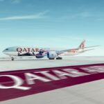 En primer plano aparecen las palabras Qatar Airways pintadas sobre el suelo, en letras blancas y fondo carmesí. Al fondo, sobre una pista, se ve un avión de esa aerolínea.