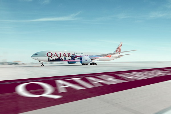 En primer plano aparecen las palabras Qatar Airways pintadas sobre el suelo, en letras blancas y fondo carmesí. Al fondo, sobre una pista, se ve un avión de esa aerolínea.
