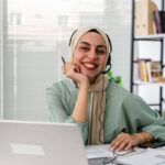 Mujer sonríe sentada ante un escritorio y una laptop, en un ambiente de oficina, mientras usa una diadema con audífono y micrófono.