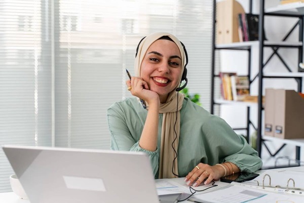 Mujer sonríe sentada ante un escritorio y una laptop, en un ambiente de oficina, mientras usa una diadema con audífono y micrófono.