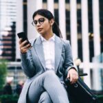 Mujer joven de negocios, con vestido ejecutivo gris y anteojos, revisa el celular mientras espera sentada, en el exterior, con una maleta pequeña.