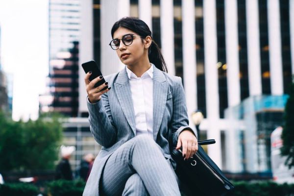 Mujer joven de negocios, con vestido ejecutivo gris y anteojos, revisa el celular mientras espera sentada, en el exterior, con una maleta pequeña.