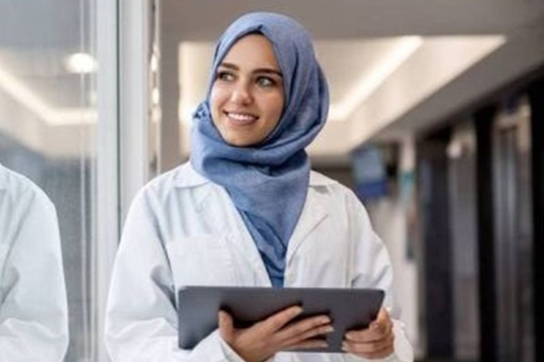 Mujer joven sonríe mientras sostiene una tablet en su mano. Usa una bata blanca de médico y un hiyab de color azul.