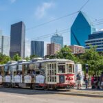 Un trolebús se acerca a una estación del McKinney Avenue Trolley, en Dallas, Texas. Al fondo, se ven varios edificios de la ciudad.