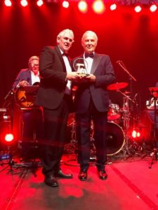 Twee mannen met prijs op podium, rode verlichting.