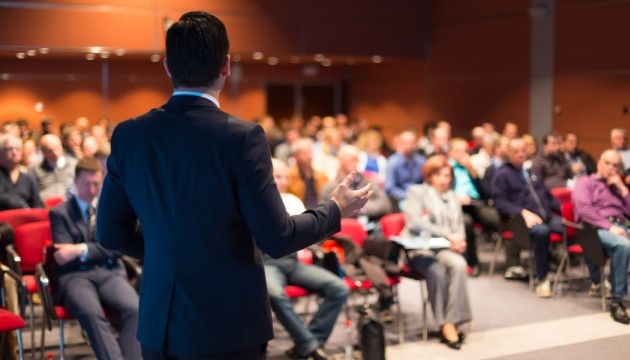 Spreker presenteert voor groot publiek in conferentieruimte.