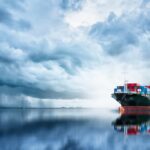 Cargo ship on reflective sea under stormy clouds.