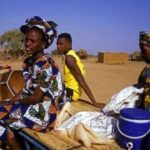 Drie mensen op kar getrokken door ezel in savanne.