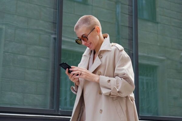 Een goed geklede vrouw met een getinte bril controleert haar telefoon terwijl ze over straat loopt.