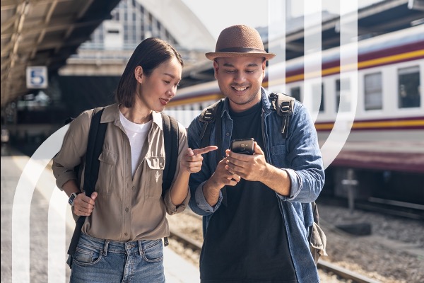 Two travelers check a smartphone beside a train platform.