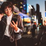 cheerful pretty young woman in cool eyeglasses and trendy wear walking on metropolis street with night lights enjoying audio songs from playlist in earphones connected to smartphone device