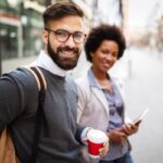 Two people smiling outdoors with coffee and tablet.