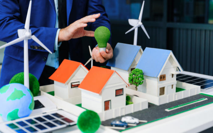a person in a business suit is holding a model of a house with solar panels and a wind turbine in front of it