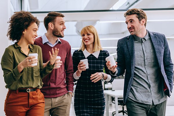 picture with a group of four people shown in front, from left to right women, women, men, each holding a cup in left hand, walking in an office environment.